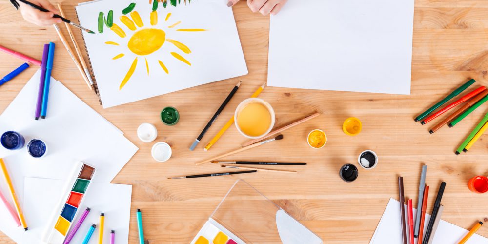 Top view hands of mother and son painting with watercolor paints together on the table